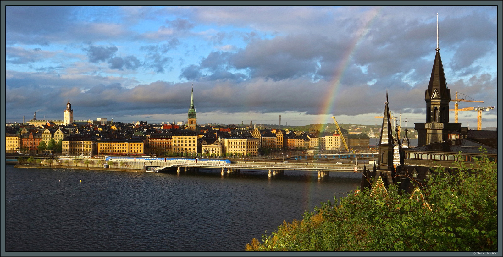 Nach einem kräftigen Regenguss bildet sich am 17.05.2023 im Licht der Abendsonne ein Regenbogen über der Stockholmer Altstadt. Auf der Brücke über den Riddarfjärden begegnen sich ein Doppelstocktriebwagen ER1 der MÄLAB als Mälartåg und ein Metrozug der Reihe C20.