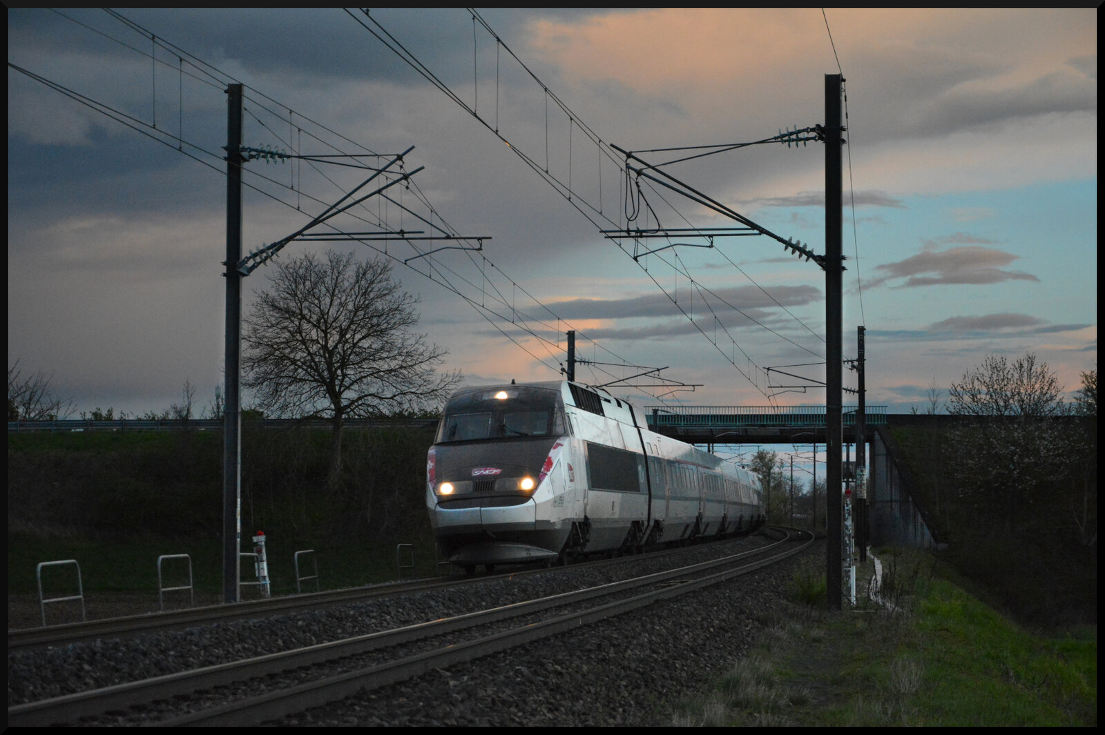 TGV 2204 Montpellier Saint-Roch - Strasbourg & TGV 5500 Montpellier Saint-Roch - Metz Ville kommen am 01.04.2024 kurz nach Sonnenuntergang bei Ostheim (Oschta) im Elsass mit 220 km/h unter der Straßenbrücke hervor.
