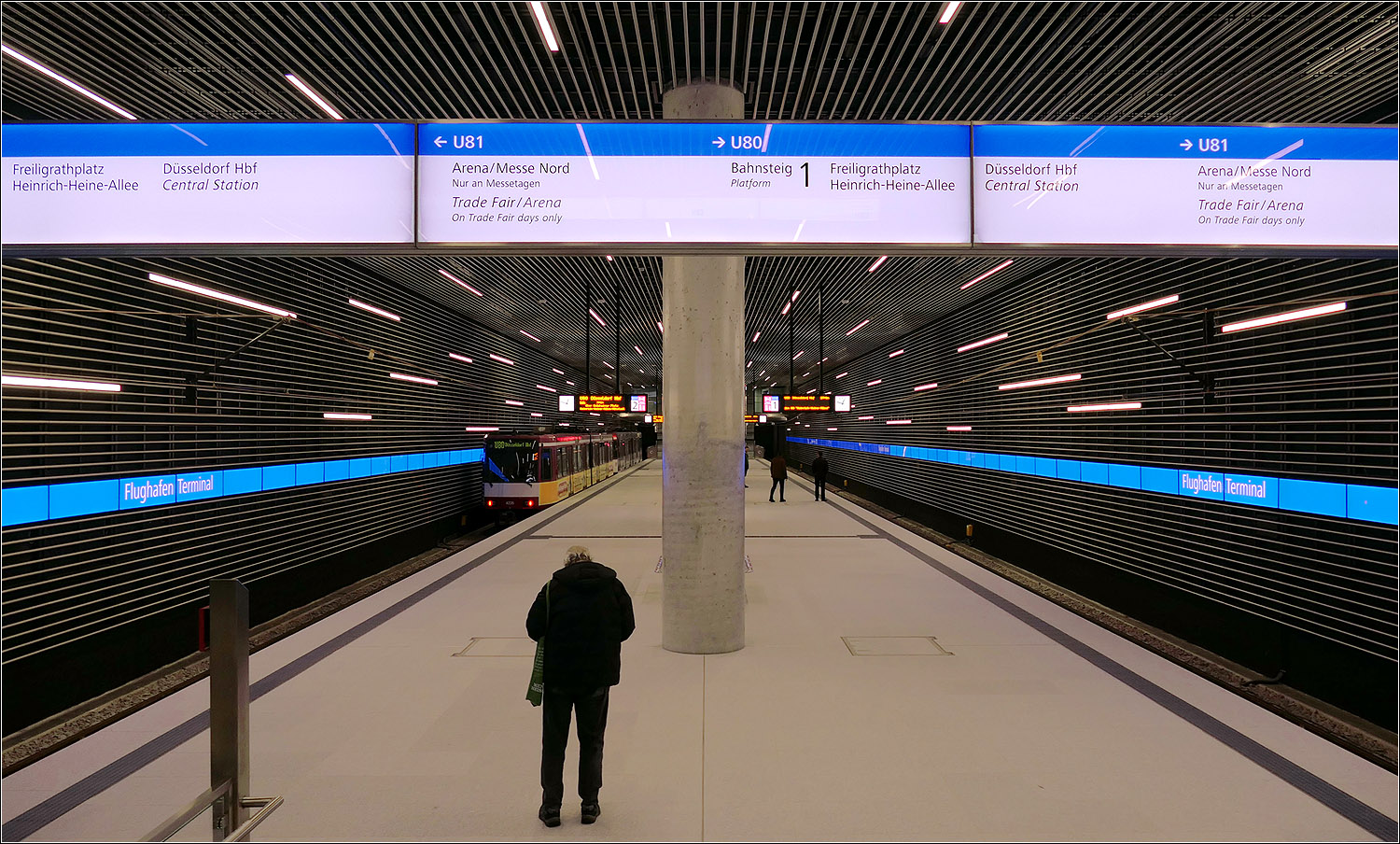 U-Station D�sseldorf Flughafen Terminal - 

Blick vom westlichen Ende der Station auf den Mittelbahnsteig in der hohen Bahnsteighalle.

Auff�llig der hellblaue Streifen an den Hintergleisw�nden. Die Anzahl der sichtbaren Linien in der Architektur d�rften mit die h�chste in einem U-Bahnhof sein. Die Lamellen ziehen sich von den W�nden �ber die Decke. Die Leuchtr�hren sind frei in den W�nden und der Decke verteilt. Fl�chtig erinnert diese Verteilung an die neuen U-Stationen in Karlsruhe, dort sind sind sie allerdings von der Decke abgeh�ngt im Raum verteilt.

12.10.2026 (M)