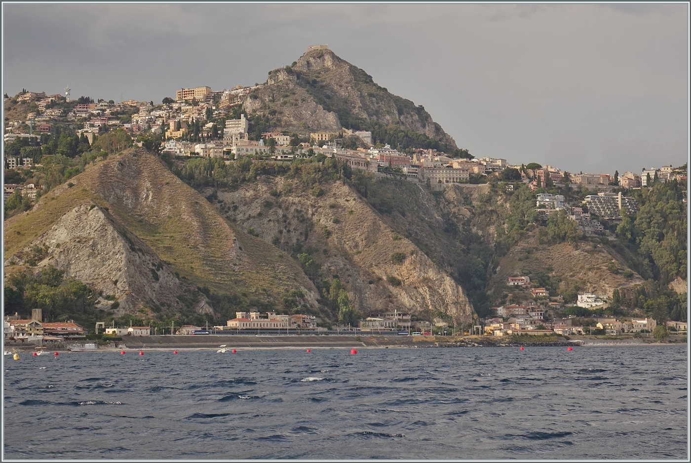 Vom Meer her ein Blick auf Tarormina und seinen Bahnhof Taormina-Giardini. Im Bahnhof stehet, soweit erkennbar,  ein langer FS Treni Turistici Italiani Zug mit je einer E 464 an der Spite und am Schluss. 

24. September 2025