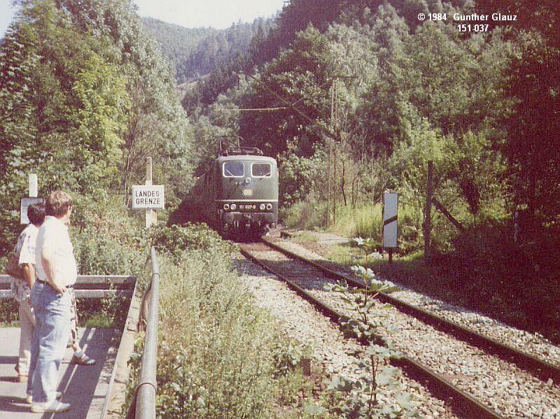 151 037 mit D-Zug Berlin - Mnchen berquert im Sommer 1984 die Grenze DDR / BRD zwischen Probstzella und Ludwigstadt. Wegen den Steigungen der Frankenbahn wurden meistens auch fr D-Zge Gterzugloks verwendet, Lokwechsel war in Prostzella, von dort war die Strecke ber die Grenze elektrifiziert. Der DDR-Grenzpfahl steht direkt hinter der Signalbarke.
