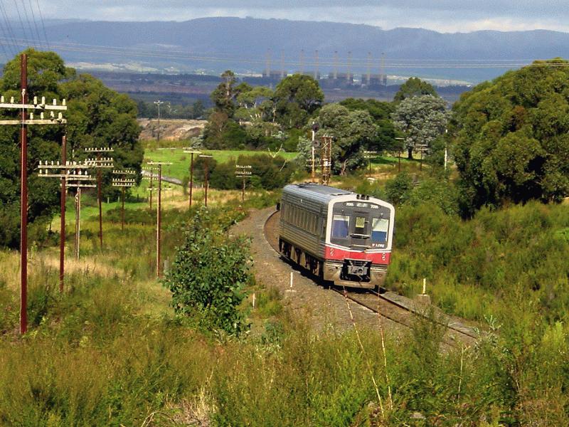 7025 der V-Line unterwegs von Morewell Richtung Moe, im Hintergrund die 4 Blcke der 1600MW Hazelwood Power Station, La Trobe Valley, April 2002
