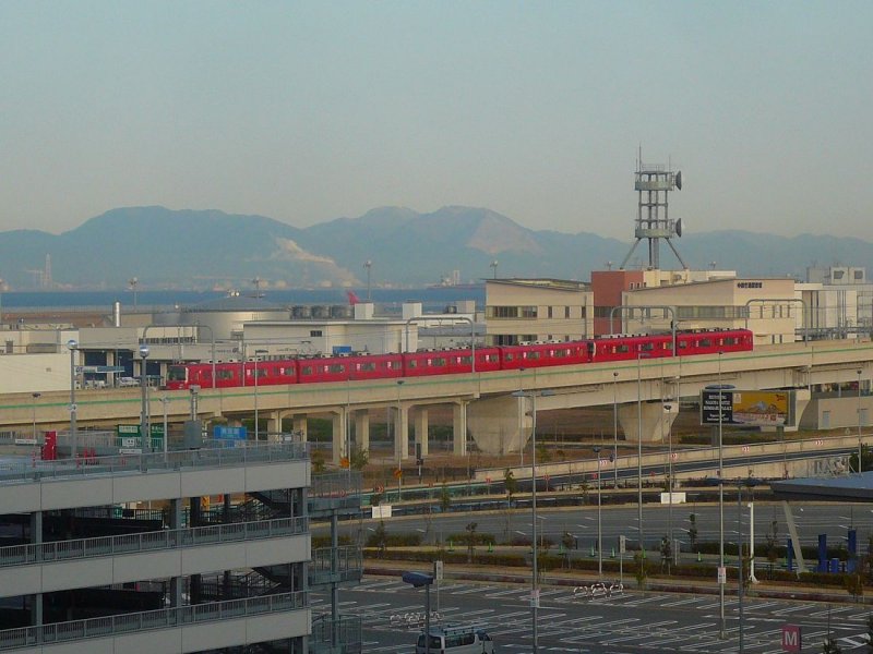 Der Meitetsu-Konzern. Ein 2+4-Wagenzug verlässt den Flugenhafen Nagoya Richtung Stadt. Im Hintergrund liegt die Bucht von Ise, dahinter das wuchtige Industriegebiet von Yokkaichi. In der Ferne erhebt sich das Suzuka-Gebirge. 5.März 2009. 