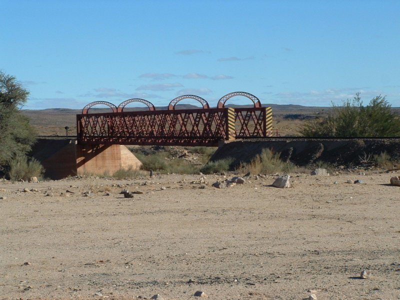 Die flache Steppe wird von einigen Trockenflusslufen durchzogen. Ein solches Rivier ist auch der Holoog, der mit einer Brcke berspannt wird. Kurz danach, hinter dem rechten Bildrand befindet sich die verwaiste Station Holoog. Einst ein wichtiger Verladepunkt fr die Farmer und Wasserstation fr die Loks. Verbindungsbahn nach Sdafrika.