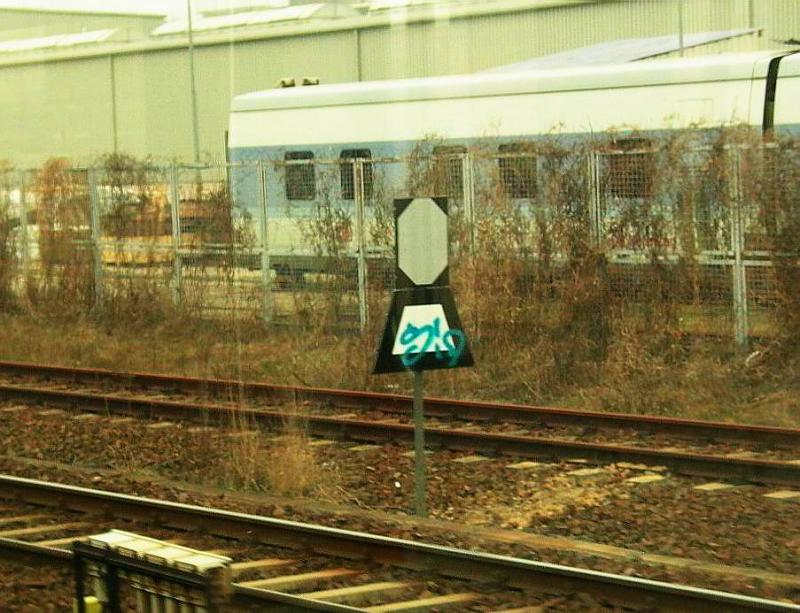 Die wahrscheinlich einzige Trapeztafel im Innenstadtbereich von Berlin. Sie ersetzt seit einigen Jahren das Einfahrsignal f�r den verbliebenen Rest des Wriezener Bahnhofs. Der Bahnhof, dessen einzige Funktion nach R�ckbau fast aller Anlagen die Bedienung des AW ist, besteht nur noch aus einem einzelnen Einfahrgleis, das an dieser Trapeztafel beginnt und an einer H-Tafel endet. Von dort aus fahren die �bergabefahrten als Rangierfahrt weiter. Interessant ist die sicher sehr seltene Kombination mit einer Eckentafel. Zu bem�ngeln ist, dass die gem�� DV 301 vorgeschriebene schwarz und wei� schr�g gestreifte Farbgebung des Pfahls fehlt.