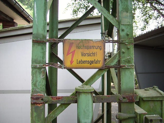 Ein sehr verdrecktes Schild in Maintal Ost das vor der Hochspannung der Oberleitung warnt.