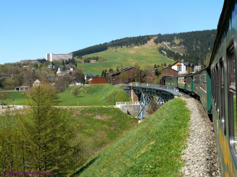 Einfahrt nach Oberwiesenthal. Die 99-772 der SDG-Fichtelbergbahn berfhrt mit ihrem Zug SDG2011 gerade den 110 m langen und 23 m hohen Httenbachviadukt hinter dem dem sich der namensgebende Fichtelberg, mit 1215 m der hchste Berg Sachsens, zeigt.
Die beiden groen holzverkleideten Gebude hinter der Lok beherbergen die Werkstatt der Fichtelbergbahn.
11.05.2008
