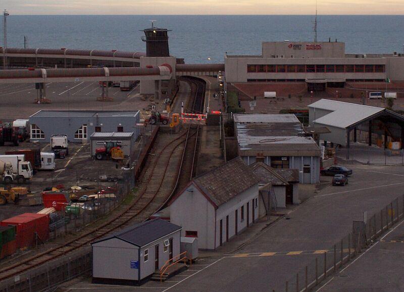 Hafen-Bahnhof Rosslare Europort am 17.02.2005 in der Abenddmmerung, er besteht nur aus einen Bahnsteiggleis und liegt direkt links neben dem Fhrgebude. Fhren verkehren von hier nach England und Frankreich. Im Bahnsteigbereich ein Bahnbergang mit Schwenk-Schranke, der Bahnbergang ist offen. Diese Schranken sieht man in Irland noch fters.