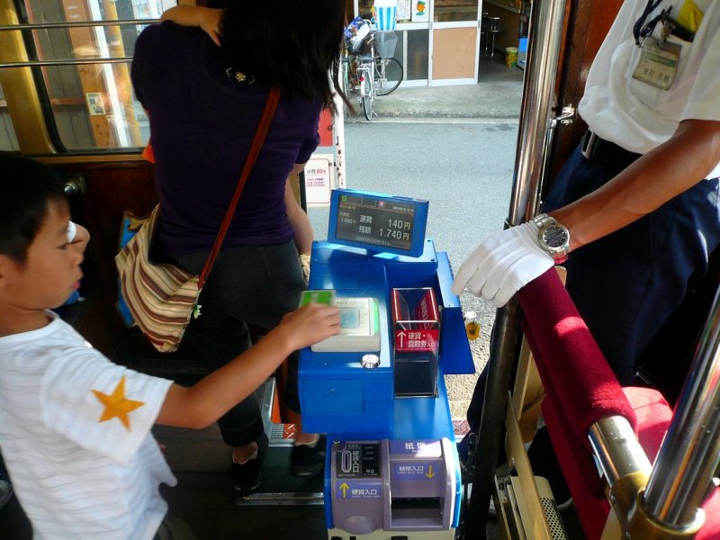 Matsuyama Strassenbahn, Serie 51-61: Ein Junge legt im Wagen 58 seine Karte auf ein Lesegert, das in Sekundenschnelle das Fahrgeld abbucht; der Vorgang und der Restbetrag der Karte werden auf dem kleinen Bildschirm angezeigt. Man kann auch bar zahlen durch Einwurf von Mnzen. Vorne sind die Schlitze zu erkennen, um Mnzen (links) oder Geldscheine (rechts) zu wechseln. Der Fahrer steht bei jeder Haltestelle auf, um den Zahlvorgang zu berwachen, wo ntig zu helfen und jeden Fahrgast einzeln zu verabschieden. 18.September 2009. 