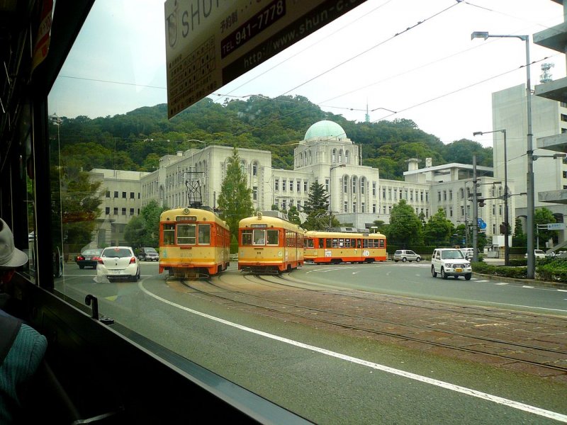 Matsuyama Strassenbahn, Serie 51-61: Vor den Verwaltungsgebuden der Prfektur kreuzen sich der herkommende Wagen 55 und die davonfahrenden Wagen 52 und (aus einer anderen Serie) 2003. Aufnahme aus einem Bus, 18.September 2009. 