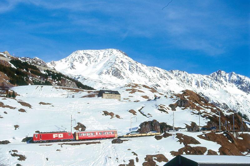 MGB FO-AUTOZUG 4353 von Sedrun nach Andermatt am 15.02.1998 Einfahrt Andermatt mit Zahnrad-E-Lok HGe 4/4II 105 - RhB B 2213 - Sklv 4833 - Rvw 4798 - Sklv 4834 - Sklv 4832. Hinweis: FO-Lok noch in alter Lackierung.
