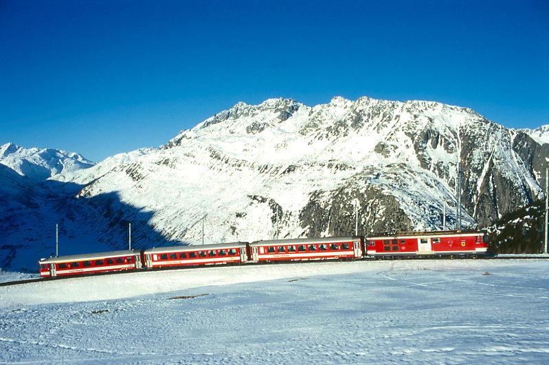 MGB FO-DIENSTZUG 6821 von Oberalppass-Calmot nach Andermatt am 15.02.1998 etwa 400m unterhalb von Ntschen mit Steuerwagen voraus ABt 4155 - B 4255 - B 4276 - Zahnrad-Gepcktriebwagen BDeh 4/4I 55. Hinweis: Fahrzeuge noch in alter Lackierung. Der abgelichtete Dienstzug ist die Rckleistung eines Wintersportzuges der die Wintersportgebiete Ntschen und Oberalppass von Andermatt aus bedient. Der Zug schlngelt sich hier bremsend in einen 4193m langen Zahnstangenabschitt mit 110 Promille Geflle in mehreren Serpentinen von Ntschen nach Andermatt im Urserental hinab. Auch am spten Vormittag ist im ganzen Tal noch Schatten bis zum 2431m hohen Furkapass, der ber der Triebwagenspitze erkennbar ist. 

