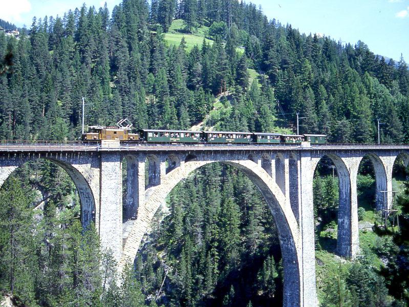 RhB FILISUR BAHNFEST Extrazug 3137 von Davos-Platz nach Filisur vom 04.07.1999 mit E-Lok Ge 6/6I 414 - B 2245 - B 2245 - B 2060 - C 2013 - D 4052II. Hinweis: Blick von Sden, Normalansicht. In den 30er-Jahren verkehrten solche Kompos als Personenzug. Allerdings stammen diese Vierachser von der Arosabahn! Tip fr Fotografen: Bei Mitfahrt bei einer der angebotenen Davos-Rundfahrten von Landquart ber Davos nach Filisur wird hier meist ein Fotohalt eingelegt. Man verlt den Zug im Bahnhof Wiesen, berquert am Zugende beim Bahnbergang die Gleise und luft dann den Wanderweg entlang Richtung Viadukt und berquert diesen. Nach etwa 8 Minuten ist dieser Fotostandpunkt erreicht. Der Zug hlt nach berquerung kurz an (mit Begleitpersonal vorher absprechen) und nimmt die Fotografen und Filmer wieder auf. Auch Autobegleiter knnen zwischen Davos und Filisur kaum zustzlich Fotos schieen, da die Anfahrt und Fuweg normalerweise zu lange dauert.