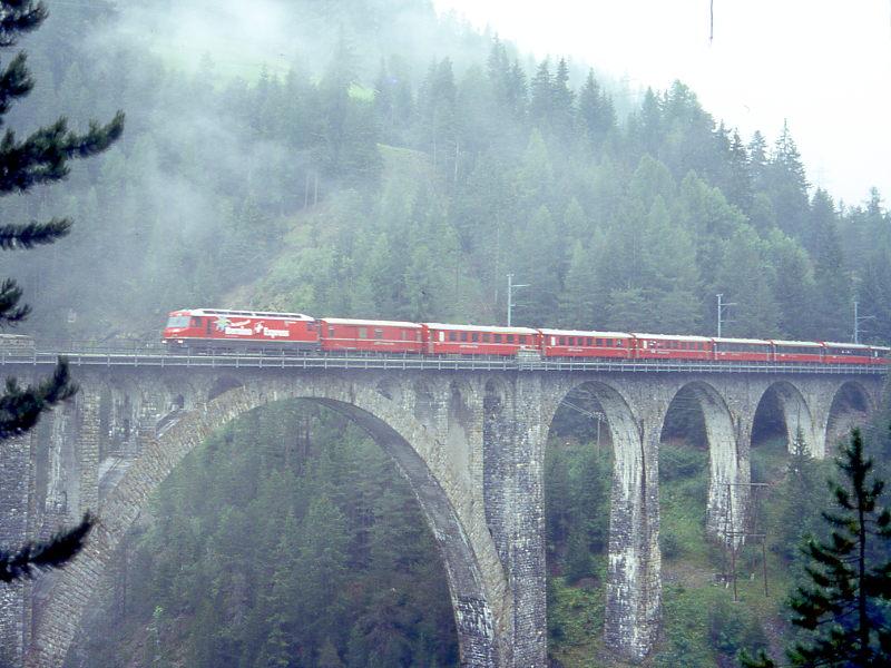 RhB Heidiland-Bernina-Expre G 503 von Landquart ber Davos-Platz - Filisur - Pontresina nach Tirano vom 30.08.1996 auf Wiesener Viadukt mit Ge 4/4III - AB 1565 - AB 1568 - AB 1567 - D 4210 - B 2468 - B 2494 - A 1271 - B 2463. Hinweis: Aufnahme bei sehr schlechten Lichtverhltnissen mit 1/15 Sekunde. Belichtungszeit und Blende 3,5 auf 100er-Diafilm. Deutlich sind die brennenden Scheinwerfer der Lok sichtbar. Erkennen Sie die Besonderheit dieses Zuges? Zusammenstellung mit passenden Werbelok extra fr diese Fotofahrt. Die RhB zeigte sich wegen des schlechten Wetters von ihrer besten Seite und spannte am nchsten Tag gleich nochmals diese Werbelok vor den Heidiland-Bernina-Expre. Sdansicht vom leicht erreichbaren und gesicherten Fotostandpunkt.