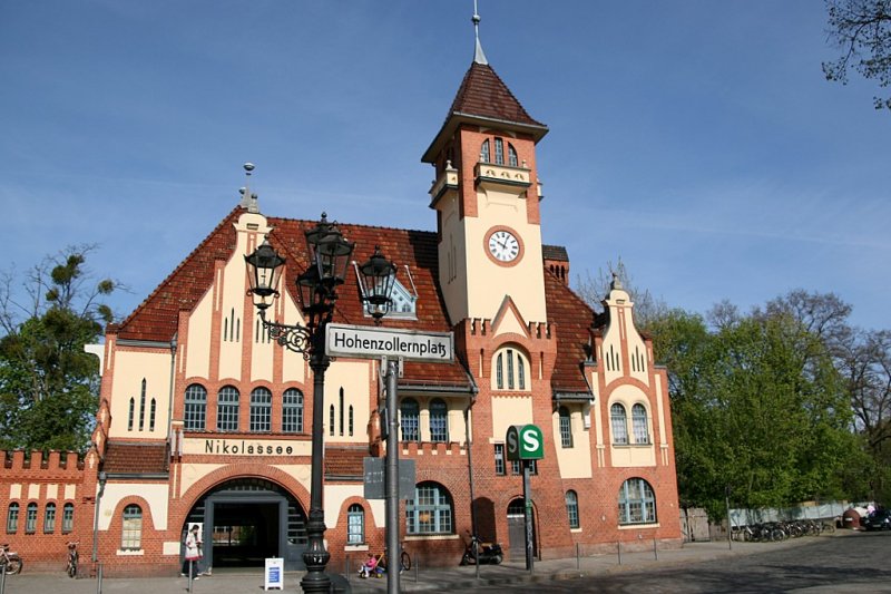 S-Bahnhof Nikolassee: Postkartenmotiv der S-Bahn. Gebaut von Brhning und Vogler 1901-02 in neogotischen Stilformen die sich mit ihrer aufwndigen Fassadengestaltung perfekt in die umliegende Villengegend anpasst. Zahlreiche Giebel Vorbauten und Zinnen zieren das Gebude mit dem markanten Turm, mit seinem kleinen Balkon.