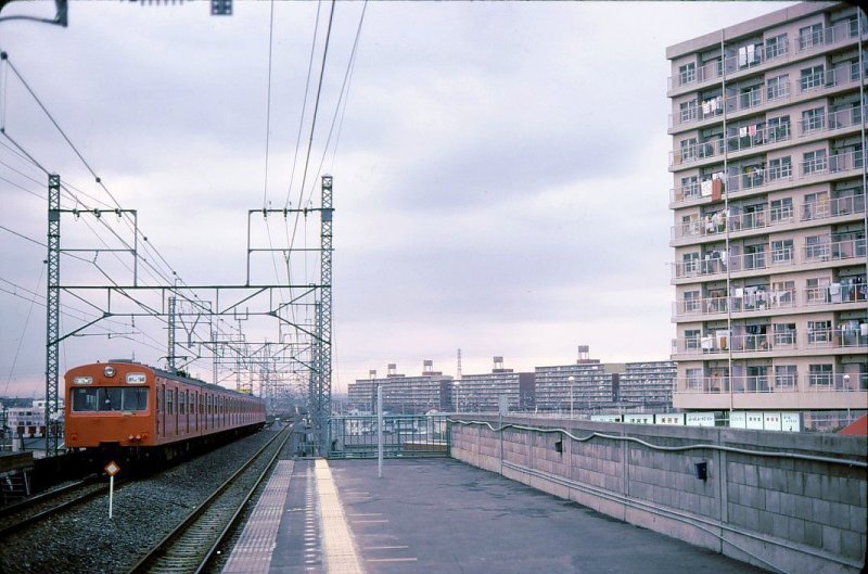 Serie 101: Ab 1973 wurden 15 6-Wagenzüge für Betrieb in langen Tunnels umgebaut (verbesserter Feuerschutz) und auf die neue, äussere Ringlinie um Tokyo (Musashino-Linie) versetzt. Statt der hübschen, kleinen Häuser, die für Tokyo typisch sind, entstanden hier draussen riesige Wohnbausiedlungen, wie im Bild sichtbar. Zug mit Frontwagen KUMOHA 101-1008 in Shin Matsudo, 31.Januar 1980. MUSASHINO-LINIE