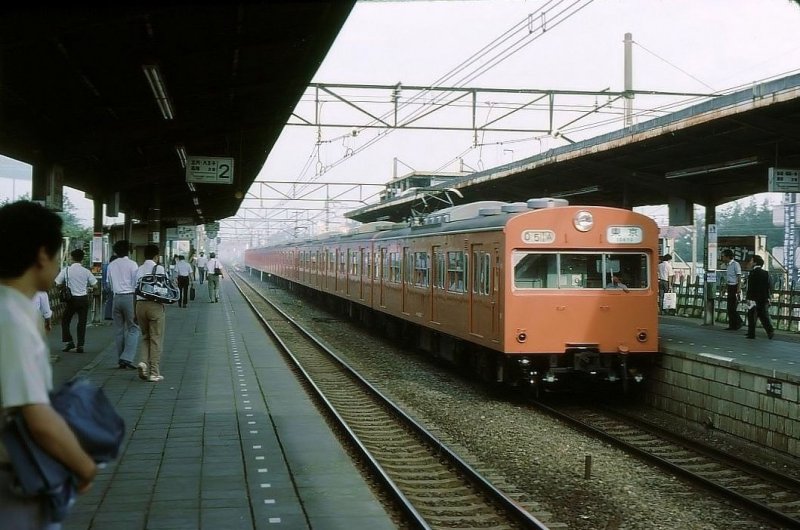 Serie 103: Nach Versuchen ab 1970 begann ab 1973 die Ausrüstung eines Teils der Wagen mit Kühlaggregaten (auf dem Dach sichtbar). Im Bild kann die stickig-heisse und unerträglich schwüle Luft im Dunst hinter dem Bahnhof erahnt werden. Im Bild ein oranger Zug der Chûô-Linie (von Tokyo nach Westen) mit Frontwagen KUMOHA 103-57 in Tokyo-Higashi Koganei, 2.Juli 1979. 