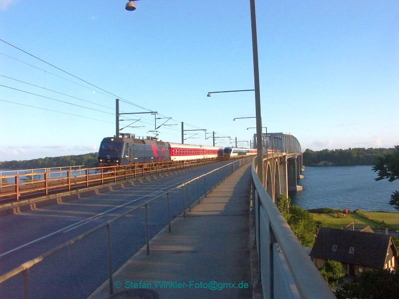 Sommer 2008 in Middelfart. Der Nachtzug aus Kopenhagen, der bis Mnchen luft, berquert gerade um 21.10 die Meerenge des kleinen Belt von Fnen herber und erreicht gerade das Ufer in Jtland. Hinten fhrt noch ein Desiro nach Fyn. Es war relativ hektisch, von hinten kamen Autos.... Leider verzeichnet mein Weitwinkel etwas, so neigen sich die beiden Masten recht und hinter der Lok zeueinander. Ich wollte unbedingt zeigen wie hoch ber dem Meer man sich hier befindet, siehe Haus rechts unten. Hoffe das Bild gefllt!
