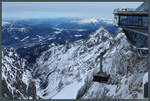 Die am 21.12.2017 in Betrieb genommene Seilbahn Zugspitze verkehrt zwischen dem Eibsee und dem Zugspitzgipfel. Sie ersetzte die auf gleicher Strecke verkehrende Eibsee-Seilbahn (Baujahr 1963). Zu sehen ist eine Gondel nahe an der Bergstation. Links im Tal die Stadt Garmisch-Partenkirchen. (Zugspitze, 03.03.2018)