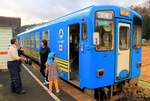 Auf der Akita Nairiku Lokalbahn im äussersten Nordwesten von Japan wartet Triebwagen 8807 eine Kreuzung ab. In dieser Zeit bringen ein Bauer und seine Frau mit grossem Vergnügen Lunch-Pakete zur Bahn, in der eine Schulklasse sitzt. Kami Hinokinai,15.November 2017.  