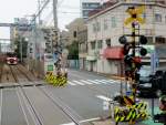 Die Stammstrecke des Keihin Kyûkô (Keikyû)-Konzerns: Am selben Ort wie das Bild mit dem blauen Zug 2157 in Hatchô Nawate, Stadt Kawasaki. Blick auf die Bahnschrankenanlage; bei allen Bahnschranken in Japan wird mit Pfeilen die Richtung angegeben, aus der sich der Zug nähert - hier im Bild leuchten beide Pfeile. Der Gegenzug ist der Achtwagenzug 1613 (Serie 1500). 25.Juni 2011.  