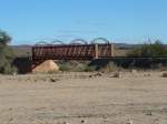 Die flache Steppe wird von einigen Trockenflusslufen durchzogen. Ein solches Rivier ist auch der Holoog, der mit einer Brcke berspannt wird. Kurz danach, hinter dem rechten Bildrand befindet sich die verwaiste Station Holoog. Einst ein wichtiger Verladepunkt fr die Farmer und Wasserstation fr die Loks. Verbindungsbahn nach Sdafrika.