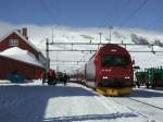 El18 2258 ist mit dem Express aus Bergen in Finse eingetroffen. Der Wintersportort in der Hardangervidda hat keine Straenverbindung. Der Versorgung luft einzig ber die Bergensbanen. Die Polizei patroliert in den Osterferien mit Schneescootern. Das einzige gleisungebundene Radfahrzeug ist ein groer Schaufelradlader, der den Bahnsteig berumt.