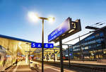 Bahnhofsschild von Salzburg Hbf, mit dem taghell beleuchteten Bahnhof dahinter.
