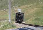 Zum Jubil�um 100 Jahre FO fuhr morgens ein Extrazug von Realp nach Andermatt um die G�ste abzuholen. Hier zwischen Realp und Hospental am 04.07.2026 im alpinen Landwirtschaftsgebiet. Andermatt hat keine Drehscheibe, daher zieht die Lok in R�ckw�rtsfahrt und passiert hier den Bahn�bergang.