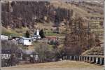 Der R1933 mit der Ge 4/4 II 633  Zuoz  umrundet die Kirche von Lavin. Die Panzersperren, auch Toblerone genannt, rechts im Bild sind noch ein Überbleibsel aus dem 2. Weltkrieg. (10.04.2015)