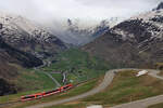 Matterhorn-Gotthardbahn Triebwagen ABDeh4/8 2028 schlängelt sich zum Oberalppass herauf, unterhalb Nätschen. Blick Richrtung Furka-Pass und in den feinen Sprühregen im Urserental. Auch hier wieder die eigentümlich gelbe Färbung des Schnees, als Folge von Sahara-Sand. 6.Mai 2024  
