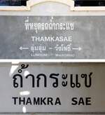 Zwischen diesen beiden Stationsschilder der Tham Krasae Station liegt das Streckengleis und ca. 6 Meter Luftlinie. - Da in der thailändischen Schrift einzelne Wörter zusammengeschrieben und es nur zum Satzende ein Zwischenraum gibt kommt es nicht selten zu unterschiedlichen Übersetzungen, welche sich in den beiden Stationsschilder widerspiegeln. - Im oberen Bild steht in der ersten Zeile  Saphan Tham Krasae  (diese Station ist vom Stationsschild 1,8 Kilometer entfernt) und in der zweiten Zeile Tham Krasae ohne Abstand und ohne dem Buchstaben  R . - Im unterem Bild befindet sich die Worttrennung an der falschen Stelle. - Bilder vom 30.November 2025.