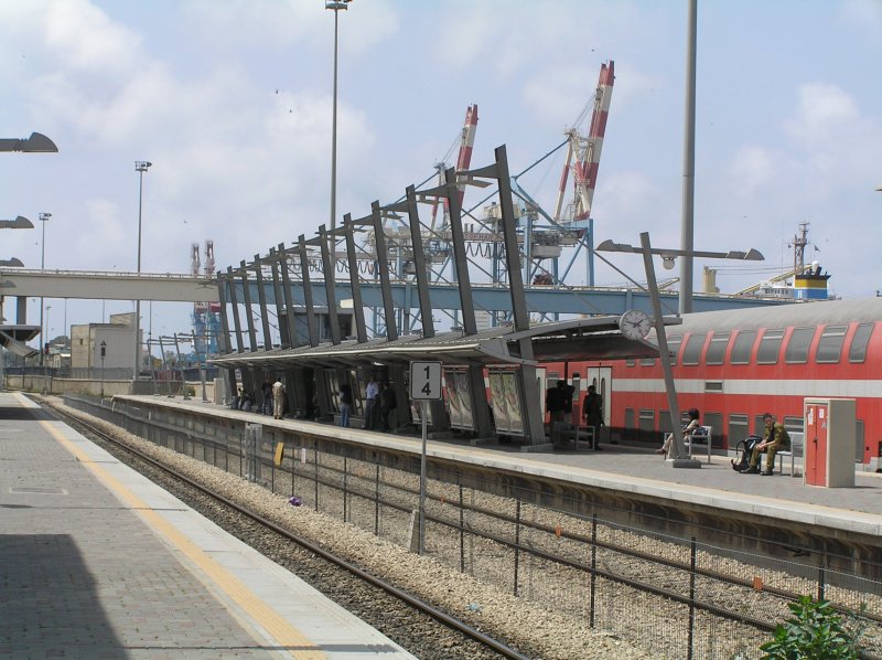 ber den am Bahnsteig 3 des Bahnhofs  Haifa Merkaz  eingefahrenen Regionalzug sind Hafenkrane und auch die Aufbauten eines Seeschiffes im Hafen Haifa zu erkennen. Bis zur Kaikante sind es vom Bahnsteig aus keine 150 m. Die berfhrung im Hintergrund ist fr die Frderbnder zum Getreidespeicher auf der anderen Gleisseite. Haifa 13.05.2007