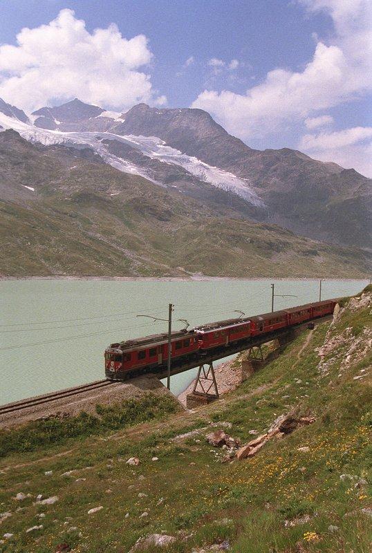 Vor majesttischer Bergkulisse berquert die Berninabahn am Lago Bianco auf ber 2200 Metern ber Meer diese Brcke. Vor der Brcke ist noch der alte Streckenverlauf zu sehen. August 1990. Gescanntes Farbnegativ. 