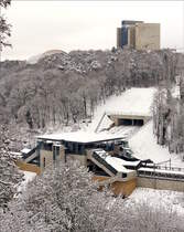Zwischen dem Boulevard Robert Schuman und dem Pont Grand-Duchesse Charlotte (Rout Br�ck) erlaubt eine L�cke im Zaun diesen Blick den neuen Bahnhof Pfaffenthal-Kircherg. 31.01.2019 (Jeanny) 

Der Bahnhof wurde auf drei Ebenen errichtet, um die 70 Meter H�henunterschied zwischen dem Quartier Pfaffenthal in der Unterstadt von Luxemburg und das Kirchberg-Plateau miteinander zu verbinden. Auf der mittleren Ebene befinden sich die Bahngleise und die Perrons. Der untere Bahnhofzugang befindet sich an der im Talgrund verlaufenden Rue Saint-Mathieu. Die Bergstation ist an die Stra�enbahnlinie angebunden. Die Gesamtanlage schafft zugleich eine Verbindung zwischen der Unterstadt und dem Kirchberg.

Der Kirchberg ist das Herz des Finanzplatzes Luxemburg und Standort mehrerer europ�ischer Institutionen (Europ�ischer Gerichtshof, Rechnungshof, Sekretariat des Europ�ischen Parlaments, Europ�ische Investitionsbank (EIB), Eurostat usw.). Zugleich ist der Kirchberg ein Wohnquartier, in dem derzeit intensiv gebaut wird. Der Kirchberg ist mit der Innenstadt durch eine Br�cke verbunden, die von allen Verkehrsmitteln und Verkehrsteilnehmern genutzt wird (Autos, �ffentlicher Nahverkehr, Velos und Fu�g�nger). Der Bahnhof umfasst zwei jeweils 250 Meter lange �berdachte Bahnsteige und eine Mittelplattform, von der die beiden parallel gef�hrten Standseilbahnen zur Bergstation auf dem Kirchberg-Plateau abfahren. Zu dieser Plattform, wie auch auf den Talgrund, gelangt man �ber Treppen, Rolltreppen und Aufz�ge. Die gesamte Infrastruktur ist f�r mobilit�tseingeschr�nkte Personen barrierefrei zug�nglich.

Die Gesamtkosten f�r die Realisierung des Bahnhofs und der Standseilbahnen beliefen sich auf 96 Millionen Euro. Das Gesamtprojekt wurde innerhalb von zweieinhalb Jahren realisiert und zum Fahrplanwechsel im Dezember 2017 in Betrieb genommen.


Da etwa 100 Meter der ungef�hr doppelt so langen schr�gen Bahnl�nge �berdeckt sind, konnte die Bahn gut in das Landschaftsbild integriert werden. Aufgrund der urbanen Nutzung wurden auch auf eine leise Fahrt wert gelegt, weshalb D�mpfungselemente auf der gesamten Strecke verbaut wurden. Auch spezielle Pumpen in der Bremshydraulik und l�rm arme L�fter und K�hlereinheiten sorgen f�r einen besonders leisen Betrieb. Das Design der Anlagen sowie die Ausstattung wurden den Bussen und Z�gen der Nationalen Gesellschaft der Luxemburgischen Eisenbahnen (CFL) nachempfunden. 