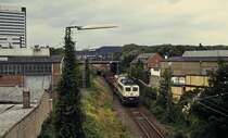 Nachtrag zur Serie von der Anschlußbedienung in Düsseldorf-Lierenfeld: Ungefähr am Endpunkt der heutigen Spitzkehre befindet sich 140 571-1, die um 1990 den Güterbahnhof Düsseldorf-Grafenberg in Richtung Derendorf verläßt. Damals wurden noch zahlreiche Anschlüsse bedient, darunter auch die Duewag (s. auch Ladung auf dem ersten Wagen hinter der Lok). Wegen des früher dichten Güterverkehrs und der oft geschlossenen Schranken wurde für ungeduldige Fussgänger ein Steg über die Gleise gebaut, von dem auch die Aufnahme entstand. Heute ist der Steg ebenso verschwunden wie der Güterbahnhof, auf dessen Gelände inzwischen Bürobauten stehen. Auch die Oberleitung wurde entfernt, denn für die einzige verbliebene Übergabe, die nur noch an Mo/Mi/Fr bei Bedarf verkehrt, genügt eine Diesellok.