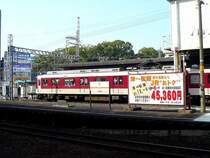 Der Kintetsu-Konzern als Konkurrenz: Mit dem Kintetsu-Zug 2811 (Typ 2800, Baujahr 1972-1979) im Hintergrund sehen wir eine grosse Tafel, auf der die JR kundtut, dass sie für Penlder in die Nachbarstadt Matsusaka billiger ist als die Kintetsu. Da der Kintetsu-Konzern aber beim Grundstückhandel die Nase vorn hat, wohnen viele Leute entlang der Kintetsu-Strecke. Tsu, 26.Februar 2009.