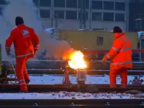 Schienenschwei�en mit der Thermit-Technik am Morgen des 14.12.2010 in Aachen West. Die Thermit-Tiegel sind Blecheimer die mit Schamott ausgekleidet sind. Darin befindet sich eine Mischung aus Eisenoxid, Aluminium und Magnesium. Mit einer Spannvorrichtung wird eine Gussform an der Schiene festgeklemmt. Darauf steht der Thermit-Tiegel der hier gerade mit einen Entz�ndungsst�bchen in Brand gesetzt wurde. Nach einigen Sekunden wird durch die chemische Reaktion eine Temperatur von ca. 2400�C ereicht, das fl�ssiges Eisen flie�t in die Form und die leichtere Aluminium-Schlacke �ber zwei �berl�ufe in Auffangschalen. Kurz abk�hlen lassen und schon kann die Form abgenommen, der �berstehende Thermit-Stahl mit einem Abscherger�t entfernt und die Naht geschliffen werden. Die Schiene ist sofort wieder belastbar. Bei den zur Zeit herrschenden Temperaturen von minus 6�C und dem eiskalten Wind scheinen die Arbeiter die abstrahlende W�rme beim verschwei�en der Schienen zu geniessen. 