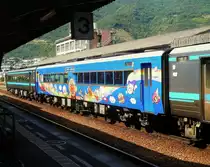 KIHA 185: Zu dieser Triebwagenserie gibt es 8 Wagen ohne Führerstand (als Serie 186). Im Bild ist KIROHA 186-2 (mit Luxusabteil RO), der mit Figuren aus der Anpanman-Geschichte dekoriert ist (Illustrationen des Zeichners Takashi Yanase (geboren 1919), der seine Jugend hier auf der Insel Shikoku verbracht hat. Anpanman erzählt von verschiedenen Brotsorten (Anpan = Brot, das mit süsser Bohnenpaste gefüllt und gross, rund und gesund ist; eine Idee, die Yanase in Hungerszeiten während und nach dem Krieg kam)). Awa Ikeda, 21.September 2009. 