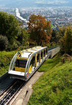 . Die neue Hungerburgbahn in Innsbruck -  Der Wagen N� 1 der neuen Hungerburgbahn erreicht am 06.10.2015 die Bergstation. (Hans)

Die Hungerburgbahn ist eine am 1. Dezember 2007 er�ffnete Standseilbahn in der Tiroler Landeshauptstadt Innsbruck. Sie verbindet die Innenstadt �ber eine geschwungene Trasse mit der Hungerburg am Berghang n�rdlich des Inns und ersetzte die alte Hungerburgbahn von 1906. Diese verlief steiler, fast geradlinig und fast genau 1 km weniger weit nur vom rechten, s�dlichen Innufer und nur auf den letzten 100 m gleichen Trasse zur selben Bergstation, woran noch die st�hlerne Innbr�cke und weit oben ein Betonviadukt erinnern. Das Projekt wurde als Public Private Partnership zwischen der Stadt Innsbruck, STRABAG und Leitner AG finanziert und realisiert. Das Unternehmen betreibt die Bahn �ber einen limitierten Zeitraum. Danach fallen die Anlagen an die Stadt zur�ck.

Die neue Hungerburgbahn gilt als eines der Wahrzeichen der Stadt, zwei Abschnitte verlaufen unterirdisch in Tunnels, sie ist eingleisig mit mittiger Ausweiche und mit 1440 mm geringf�gig breiter als in Normalspur ausgef�hrt

Die neue Hungerburgbahn verf�gt wie ihr Vorg�nger �ber 2 Fahrgastkabinen, welche sich auf der Mitte der Strecke begegnen. Der Wagen N� 1 ist gelb lackiert mit silbernen Tr�gern, Wagen N� 2 tr�gt einen blauen Lack mit ebenfalls silbernen Tr�gern. Die Wagen bestehen jeweils aus f�nf, in einem gemeinsamen Rahmen aufgeh�ngten, Fahrgastabteilen. Die aktive Verstellung der Neigung erlaubt ein Ein- und Aussteigen aus horizontalen Abteilen an den Haltepunkten. Mit den beiden jeweils 130 Personen fassenden Fahrzeugen wird eine Bef�rderungskapazit�t von 1.200 Personen pro Stunde und Richtung erreicht. Desweiteren war es aufgrund der Trassenf�hrung, die vom flachen Bereich in einen sehr steilen Bereich �bergeht, notwendig, das Seil �ber Seilrollen im Bereich der Geleise niederzuhalten. Diese Neuentwicklung ist weltweit erstmals auf dieser Anlage eingesetzt worden.

Die technischen Daten der neuen Hungerburgbahn:

H�he Talstation: 569 m
H�he Bergstation: 857 m
H�henunterschied: 288 m
Fahrzeit: 8:10 min
Betriebsgeschwindigkeit: 10 m/s
Wagenfassungsraum: 130 Pers.
F�rderleistung: 1200 P/h
Streckenl�nge: 1,838 km
Spurweite: 1440 mm
