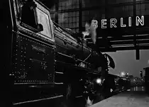 Eine Abwandlung des Filmtitels  Berlin – Die Sinfonie der Großstadt . Einer der experimentellen Dokumentarfilme ( Walther Ruttmann, im September 1927 in Berlin uraufgeführt), den ein Eissenbahnfreund wohl kennt, und der ansonsten auch jedes Fotografenherz erfreut. 
03 2030 steht vor D 1395, bald geht es los auf die regennasse Strecke. 
Kein Stativ dabei, da muss eine Belichtungsreihe 1/15 , 1/8 und 1/4 bei Offenblende helfen. Wo ist am meisten von der Lok zu erkennen - und wo habe ich am wenigsten gewackelt. Die Nietenreihe im harten Licht der Hallenbeleuchtung und die Leuchtschrift an der Halle - das musste einfach sein. 
Dies ist ein Scan vom Negativ mit 0,25 Sekunden Belichtung, ausgerichtet etwas außerhalb der Mitte, der Schrift wegen. 
