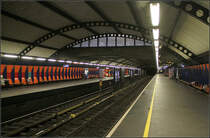 Stortinget, der Mittelpunkt des Osloer U-Bahnnetzes -

Nicht immer war das Osloer U-Bahnnetz einheitlich. Früher gab es zwei nicht kompatible Systeme im West und Osten der Stadt, die auch im Zentrum an unterschiedlichen, weit auseinander liegenden Stationen endeten. Das westliche eher eine Vorortbahn mit Oberleitungen und nicht kreuzungsfreien Strecken, einschließlich der Holmenkollenbahn endete seit 1928 unterirdisch in der Station Nationaltheatret. Das östliche Netz, eine echte U-Bahn mit Stromschienen hatte ihren Ausgangspunkt auf der anderen Seite des Zentrums am Jernbanetorget, dem Osloer Hauptbahnhof. Diese 'echte' U-Bahn wurde 1966 eröffnet nachdem vorher schon einzelne Strecken im Straßenbahnvorlauf betrieben wurde.

Es gab dann immer Pläne, die beiden Teilnetze zu verbinden, ohne dass an einen durchgehenden Betrieb gedacht wurde. 1977 wurde das Ostnetz (U-Bahn) nach Westen verlängert bis zur Station Sentrum (heute Stortinget). Dieser wurde 1983 aufgrund von Baumängeln wieder geschlossen und 1987 wiedereröffnet. Zu diesem Zeitpunkt wurden auch die Westlinien (Oberleitungsbetrieb) bis hier her verlängert und endeten in der im Bild gezeigten Bahnsteighalle. Die U-Bahn von Osten her wendete in einer Schleife um diese Bahnsteighalle herum, mit zwei getrennten Bahnsteige für ankommende und abfahrende Züge. Diese Bahnsteige sind direkt über Fußgängertunnels mit den mittleren beiden Gleisen der Westlinien verbunden. Quasi eine viergleisige Station mit zwei Mittelbahnsteigen zum direkten umstiegen in gleicher Fahrtrichtung. 

Ab 1993 wurde ein erster durchgehender Betrieb zwischen den Ost- und Westlinien eingerichtet, der nach und nach auf alle Linien übertragen wurde. Es gab dann Fahrzeuge, die sowohl für Oberleitungs- als auch Stromschienenbetrieb geeignet waren.
Inzwischen sind aber die Westlinien ebenfalls komplett auf Stromschienenbetrieb umgebaut und bis auf die Holmenkollenbahn auf kreuzungsfrei ausgebaut.

Der durchgehende Betrieb findet auf den beiden im Bild gezeigten Gleisen statt, während die Wendeschleife für Verstärkerzüge aus/in Richtung Osten genutzt wird.

Oben schneidet die Fußgängerebene mit einer Art Brücke in die gewölbte Decke der Bahnsteighalle ein.

30.12.2013 (M)