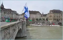 Kursschiff L�llek�nig wartet auf Passagiere und ein Combino f�hrt �ber die Mittlere Rheinbr�cke, wo unz�hlige Flaggen auf das bevorstehende Basel Tattoo hinweisen.